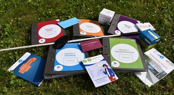 Inklusionskisten Ordner, Bücher, Hefte, ein Blindenstock und eine Augenmaske liegen auf einer Wiese.